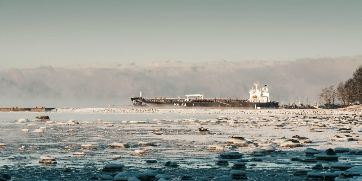 A large oil tanker navigating the narrow Strait of Hormuz, with distant coastline and calm blue water under a hazy sky.