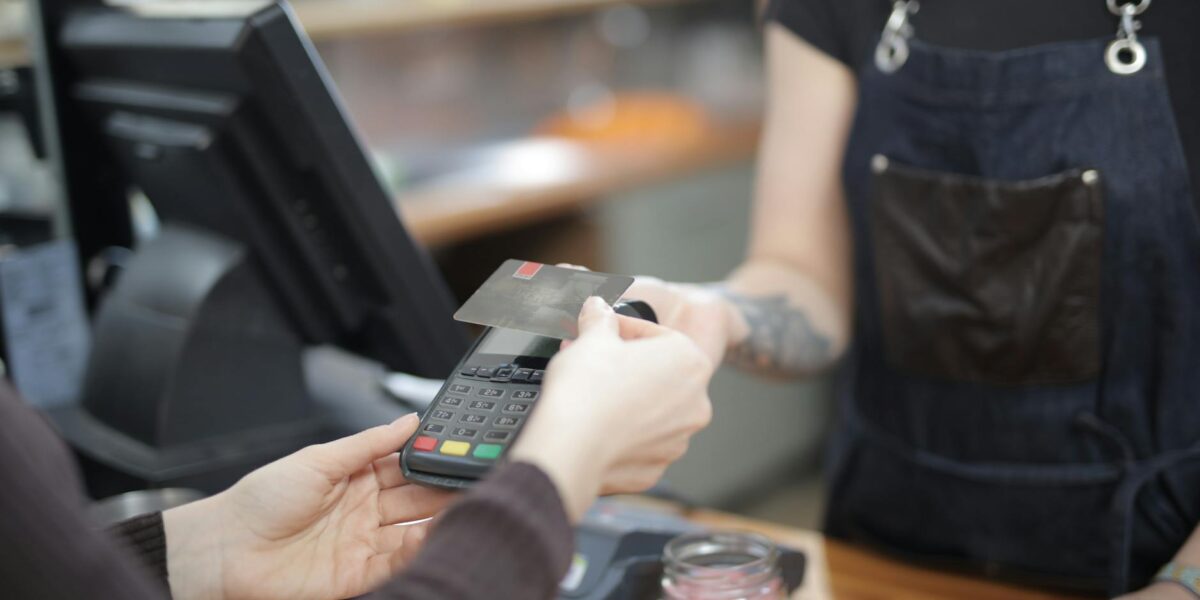 A customer's hand tapping a digital payment terminal at a cafe counter, with a tip screen visible, illustrating the spread of tipping culture overseas.