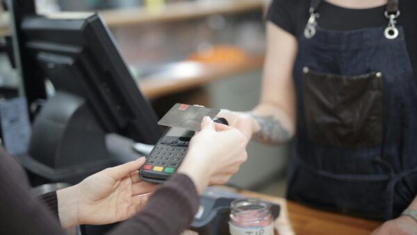 A customer's hand tapping a digital payment terminal at a cafe counter, with a tip screen visible, illustrating the spread of tipping culture overseas.